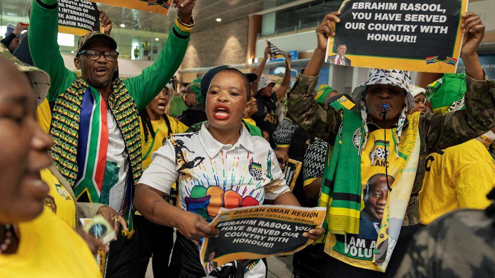 Supporters of South Africa expelled ambassador to the US Ebrahim Rasool, many dressed in the yellow, green and black colours of the ANC, wait to greet him at the Cape Town International Airport on 23 March 2025. Some hold posters reading: 'Ebrahim Rasool, you have served our country with honour!!'